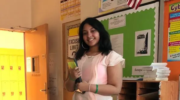 standing student that is wearing pink holding a book inside of a classroom