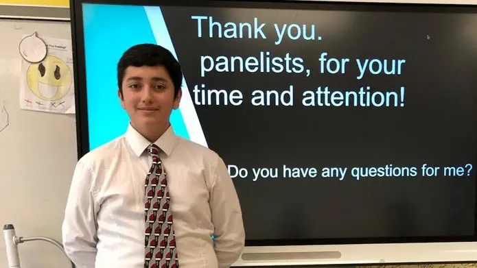 standing student in a suit and tie standing in front of a screen that says thank you panelists for your time and attention